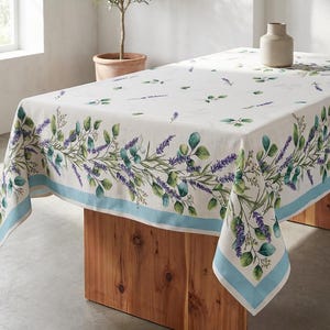 May include: A rectangular white tablecloth with a lavender and green floral border and a light blue trim. The tablecloth is draped over a wooden table. A small olive tree in a terracotta pot is in the background.