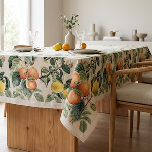 Peut inclure: Une nappe rectangulaire avec un imprimé botanique d'oranges et de citrons sur fond crème. La nappe est posée sur une table en bois, avec des chaises assorties. La scène se déroule dans une pièce lumineuse avec une fenêtre.