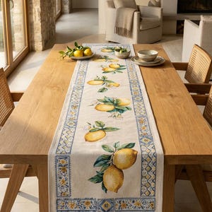 Puede incluir: Camino de mesa rectangular con diseño de limones. El corredor presenta ilustraciones de limones con hojas verdes y flores blancas sobre un fondo crema. Los bordes están adornados con un patrón geométrico azul y amarillo. Colocado sobre una mesa de madera.