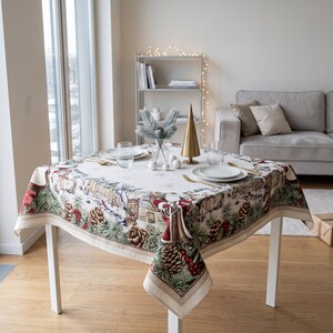 Könnte beinhalten: Eine quadratische Tischdecke mit festlichem Wintermotiv aus Tannenzapfen, immergrünen Zweigen, roten Beeren und kleinen Häusern. Der Stoff hat eine cremefarbene Mitte mit einer detaillierten Bordüre, gedeckt mit weißen Tellern und Gläsern.