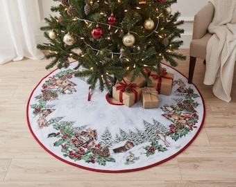 May include: A decorated Christmas tree with ornaments and lights stands above a white tree skirt. The skirt has a red border and features a winter scene with houses, trees, and animals. Wrapped gifts with red bows are placed beneath the tree.
