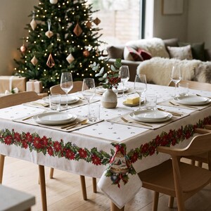 May include: A rectangular dining table covered with a white tablecloth featuring a red poinsettia and green holly leaf border. The table is set with white plates, clear glassware, gold-toned cutlery, and a central ceramic vase with greenery.