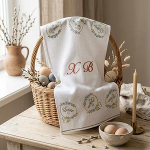 May include: A white linen towel with embroidered floral wreaths and the letters "X B" in red. The towel is draped over a wicker basket filled with Easter eggs. A ceramic bowl with eggs and a candle are on a wooden table.