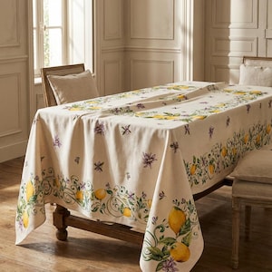 Puede incluir: Un mantel color crema con un patrón repetido de limones amarillos, hojas verdes y flores moradas. El mantel está sobre una mesa de madera, creando una pieza decorativa para comer o entretener.
