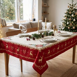 Puede incluir: Un mantel rectangular con un centro de color crema y un borde rojo decorado con patrones de renos y corazones dorados. Las esquinas muestran motivos florales, y la tela cubre una mesa de comedor de madera dispuesta con platos blancos, copas de vino y dos velas altas.