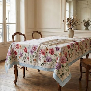 May include: A rectangular tablecloth with a floral pattern featuring red, pink, and purple flowers on a cream background. The tablecloth has a light blue scalloped border and is draped over a wooden table. The room has wooden floors and two wooden chairs.