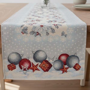 May include: A light blue Christmas table runner with a festive design. The runner features a pattern of snowflakes, holly, and ornaments in red, silver, and white. The runner is displayed on a wooden table.