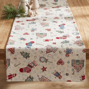 Puede incluir: Camino de mesa navideño crema con un estampado repetido de casas de jengibre, calcetines, árboles y bastones de caramelo en rojo, verde y azul. El camino de mesa está sobre una mesa de madera, con una ramita de pino y un ciervo decorativo.