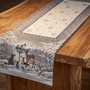 May include: A rectangular table runner with a winter forest scene, featuring deer, rabbits, and a birdhouse. The central section has a cream background with star patterns. The runner is placed on a wooden table.