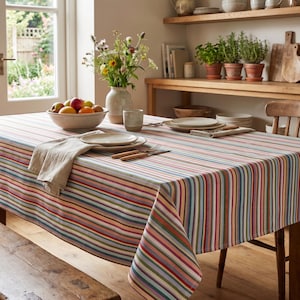 Op de afbeelding: Een rechthoekig tafelkleed met kleurrijke verticale strepen in rood, blauw, groen en oranje bedekt een houten tafel. De tafel is gedekt met borden, bestek en een beige linnen servet. Op de voorgrond staat een houten bank.