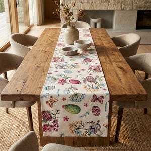 Pode incluir: Uma mesa de jantar rústica de madeira com um caminho de mesa floral com desenhos de borboletas e ovos. Cadeiras estofadas bege circundam a mesa, com um vaso de flores secas e tigelas de pedra. A mesa está posta.