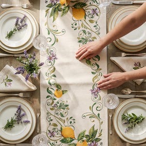 Op de afbeelding: Een tafelsetting met een crèmekleurige geborduurde tafelloper met citroenen, groene bladeren en paarse bloemen. Witte borden met gouden randen, bestek en kristallen glazen zijn rond de loper gerangschikt. Verse lavendel en citroenen zijn ook aanwezig.
