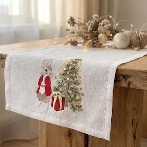Pode incluir: Um caminho de mesa de linho branco com bordados com tema natalino. O design inclui um coelho usando um casaco vermelho, uma árvore de Natal com lantejoulas douradas e uma caixa de presente vermelha. O caminho de mesa é exibido em uma mesa de madeira.