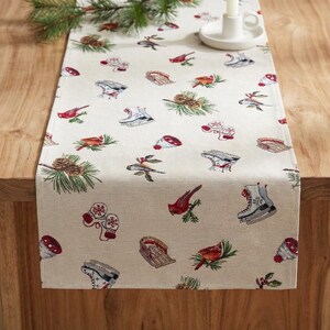 Puede incluir: Camino de mesa beige con un estampado de invierno con cardenales rojos, patines de hielo, piñas y mitones. El camino de mesa está colocado sobre una mesa de madera, con una ramita de pino y un candelabro.
