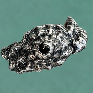 Può includere: Un anello in argento caratterizzato da una superficie organica altamente strutturata che imita l'aspetto di una conchiglia naturale o di una formazione rocciosa. Il metallo presenta una finitura lucida e riflettente con profonde fessure scure.