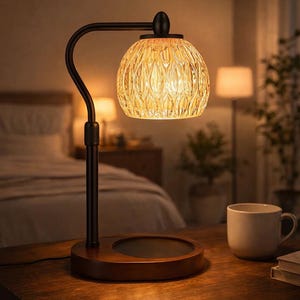 Könnte beinhalten: Dekorative Tischlampe mit warmem Licht. Sie hat ein dunkles Metallgestell und einen strukturierten, bernsteinfarbenen Glasschirm. Die Lampe steht auf einem Holzsockel. Eine weiße Tasse und Bücher befinden sich auf dem Tisch.