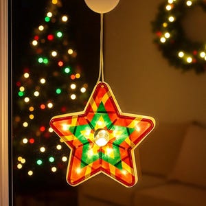 May include: A vibrant, star-shaped Christmas decoration suspended in front of a window. The star features a geometric pattern in red, green, and yellow. A small light bulb illuminates the centre, with a white disc at the top.