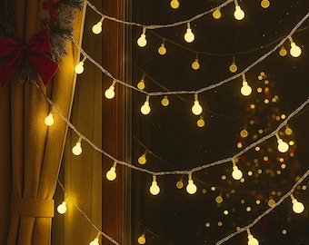 May include: Warm, glowing string lights with round bulbs are strung across a window. A Christmas wreath with a red bow hangs to the left. The lights create a cozy, festive atmosphere, perfect for holiday decor.