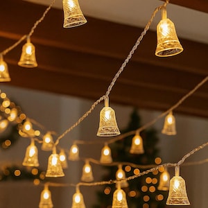 May include: String of bell-shaped fairy lights with a warm, yellow glow. The lights are strung on a thin, silver wire and are hanging from a wooden ceiling. The background is blurred, with a Christmas tree visible.
