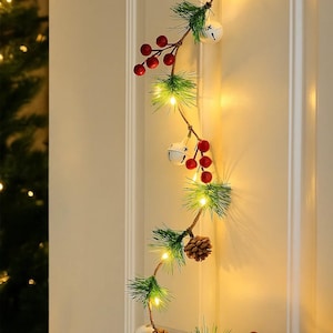 Puede incluir: Una guirnalda navideña festiva con agujas de pino artificiales, bayas rojas, campanas blancas y pequeñas luces blancas cálidas. La guirnalda también presenta piñas y está colgada en una puerta blanca. Ideal para decoración navideña.