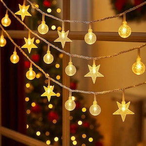 Puede incluir: Luces de cadena con bombillas en forma de estrella y globo de color amarillo cálido. Las luces están colgadas frente a una ventana, con un árbol de Navidad visible en el fondo. Las luces crean un ambiente festivo.