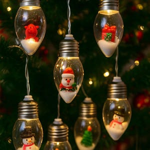 Puede incluir: Cadena de luces navideñas con adornos transparentes en forma de bombilla. Cada bombilla contiene una figura navideña en miniatura, como un reno, Papá Noel, un muñeco de nieve, un árbol de Navidad y regalos. Las luces están ensartadas en finos cables plateados.