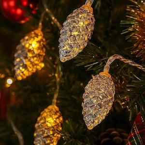Peut inclure: Guirlande lumineuse de Noël avec des ampoules en forme de pommes de pin. Les lumières dorées émettent une lueur chaleureuse. Les pommes de pin sont fixées à un fil transparent fin, sur un fond de sapin de Noël.