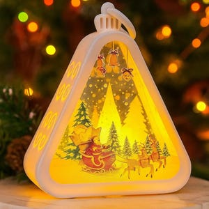 May include: A white, triangular Christmas ornament-shaped light with a festive scene inside. The scene depicts Santa Claus in a sleigh pulled by reindeer, with trees and bells. The light has a handle at the top.