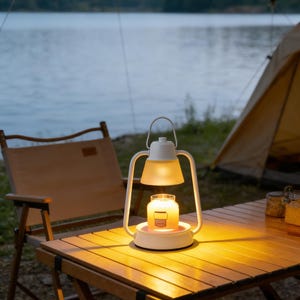 May include: A white and yellow lantern with a lit candle sits on a wooden table. The lantern has a handle and a frosted glass shade. A camping chair and tent are in the background, with a lake visible.