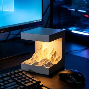 Puede incluir: Una lámpara de escritorio blanca en forma de cubo con un diseño de cordillera iluminada. La lámpara está sobre un escritorio negro junto a un teclado y un ratón. El fondo incluye un monitor de ordenador y una torre de ordenador.