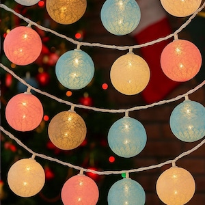 Puede incluir: Una cadena de luces decorativas con globos en varios colores, incluyendo rosa, azul, dorado y amarillo. Las luces están ensartadas en un cable blanco y están encendidas, creando un brillo cálido y acogedor. Ideal para decoraciones festivas.
