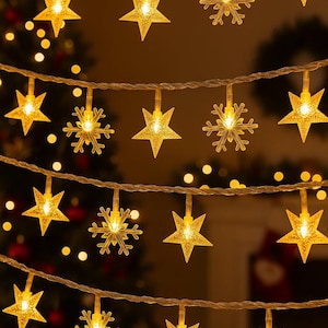 Puede incluir: Luces de cadena con formas de estrella y copo de nieve. Las luces amarillas cálidas están colgadas de un cable transparente. El fondo borroso sugiere un ambiente festivo, ideal para celebraciones navideñas.