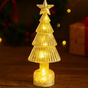 Può includere: Una piccola decorazione per albero di Natale illuminata. L'albero è realizzato in vetro trasparente strutturato e ha una stella in cima. L'albero è illuminato dall'interno, creando un caldo bagliore dorato. La base è un piccolo piedistallo rotondo.