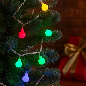 May include: A string of colorful Christmas lights with round bulbs in red, green, yellow, and blue. The lights are strung on a silver wire and are displayed on a green Christmas tree. A red gift box with a gold bow is in the background.