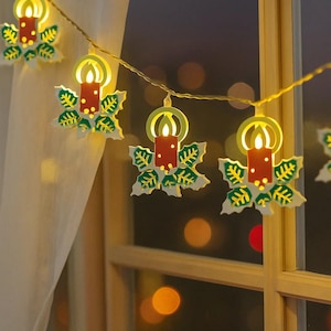 May include: A festive string of Christmas lights. Each light features a candle-shaped ornament with a red candle and a yellow flame, set within a green and white holly leaf design. The lights are strung on a thin, gold wire.