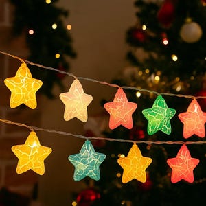 Può includere: Una stringa di luci a forma di stella in vari colori, tra cui giallo, rosa, verde e blu. Le luci sono infilate su un filo sottile e sono illuminate, creando un bagliore caldo e festoso. Lo sfondo presenta un albero di Natale.