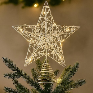 Könnte beinhalten: Ein silberner Drahtstern als Weihnachtsbaumspitze mit eingebetteten Lichtern und klaren Kristallakzenten. Der Stern ist auf einem grünen Weihnachtsbaum befestigt. Eine festliche Dekoration für die Weihnachtszeit.