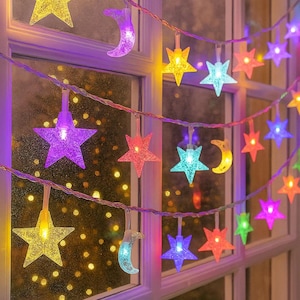 Puede incluir: Una cadena de luces decorativa con forma de estrellas y lunas crecientes multicolores. Las luces son de varios colores, incluyendo amarillo, morado, azul y rojo. Las luces cuelgan frente a una ventana, creando una exhibición festiva y decorativa.