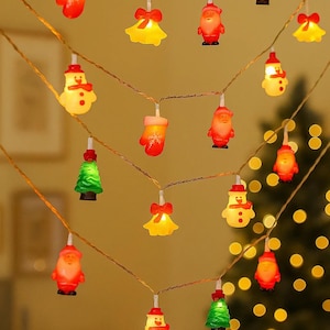 May include: Christmas string lights featuring miniature red mittens, snowmen, Santa figures, green Christmas trees, and yellow bells. The lights are strung on a thin wire, creating a festive holiday decoration.