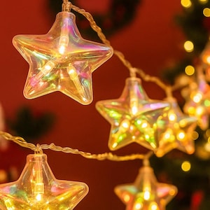 Pode incluir: Close-up de uma sequência de luzes de Natal em forma de estrela. As estrelas transparentes e iridescentes são iluminadas com luzes brancas quentes. O fundo é vermelho desfocado com uma grinalda e uma árvore de Natal.