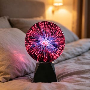 Może przedstawiać: Lampa z kulą plazmową z czarną geometryczną podstawą. Szklana kula wyświetla żywy pokaz czerwonych i niebieskich wyładowań elektrycznych. Lampa stoi na łóżku z pościelą w neutralnych odcieniach.