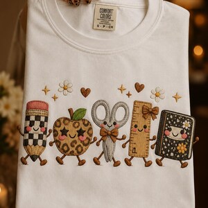 May include: A white t-shirt featuring embroidered cartoon school supplies including a checkered pencil, a leopard print apple, scissors with a bow, a wooden ruler, and a black notebook, all with smiling faces, surrounded by small flowers and hearts.