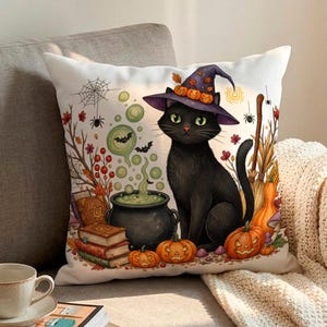 May include: A square throw pillow featuring a black cat wearing a purple witch hat adorned with small orange pumpkins. The illustration includes a bubbling green cauldron, stacked books, orange pumpkins, autumn foliage, and a spider web.