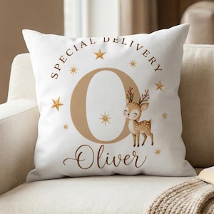 Può includere: Un cuscino quadrato bianco con una grande lettera O color marrone chiaro, un piccolo cervo dei cartoni animati e il testo Special Delivery e Oliver in marrone. Il design include diverse piccole stelle dorate sparse attorno alla lettera e al cervo.