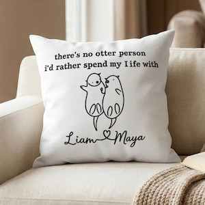 Könnte beinhalten: Ein weißes quadratisches Dekokissen mit einer schwarzen Strichzeichnung von zwei Ottern, die Händchen halten. Über den Ottern steht der Text "there's no otter person i'd rather spend my life with" und darunter die Namen "Liam" und "Maya", verbunden durch ein Herz.
