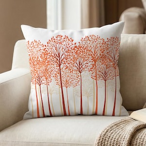 May include: White throw pillow with a repeating tree design in shades of orange and red. The trees have long, thin trunks and full, leafy crowns. The pillow is on a beige sofa.