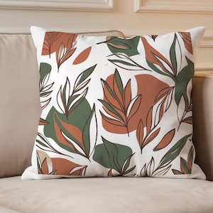 Può includere: Un cuscino quadrato con sfondo bianco e un motivo ripetitivo di foglie stilizzate e forme organiche nei toni dell'arancione terracotta, verde oliva e verde scuro. Il tessuto presenta una trama visibile su tutto il disegno.
