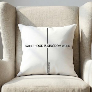 May include: A white square cushion with the text "FATHERHOOD IS KINGDOM WORK" in black, centered. A vertical black line bisects the text. The cushion is on a beige armchair. The text "PROVERBS 22:6" is below.