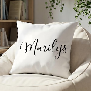 May include: A square white throw cushion featuring the name Marilys printed in a black cursive script font. The cushion has a smooth, solid white fabric cover and is positioned centrally, displaying the text clearly across the front surface.