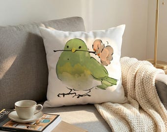 Puede incluir: Un cojín cuadrado blanco con una ilustración estilo acuarela de un pájaro verde redondo que sostiene en su pico una rama fina marrón con pequeñas flores de color beige rosado. El pájaro tiene un contorno negro simple y está centrado.